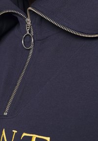 Sudadera de cuello alto azul marino con cremallera de metal, tirador circular y texto amarillo bordado en la parte delantera.