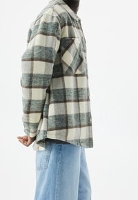 Geruite flanellen shirt in groen, wit en bruin, met een borstzak, knoopsluiting en een ontspannen pasvorm, gecombineerd met lichtblauwe denim jeans.