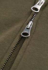 Tejido verde oliva con cremallera, de acabado texturizado, que presenta una cremallera plateada y un tirador con marca. La vista en primer plano resalta los detalles.