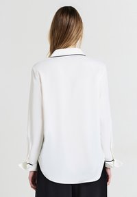 Frau mit langen Haaren, die eine weiße Bluse mit langen Ärmeln und schwarzer Einfassung am Kragen und an den Manschetten trägt, kombiniert mit schwarzen Hosen und mit dem Rücken zur Kamera steht.