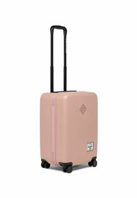 Valise rigide de couleur rose clair. Elle présente un extérieur texturé, une poignée noire rétractable et quatre roues noires pour plus de mobilité.