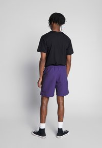 Musta lyhythihainen t-paita yhdistettynä violetteihin shortseihin. Shortseissa on joustava vyötärönauhavyö ja takataskun yksityiskohta. Jalkineina mustat korkeavartiset tennarit.