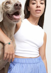 Perro gris con acentos marrones está de pie al lado de una persona que lleva una blusa sin mangas blanca y pantalones cortos a rayas azules. Se puede ver una pulsera de plata con un charm de turquesa.
