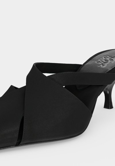 Sort mule med spids tå, krydsede remme og slank mellemhøj hæl på hvid baggrund.