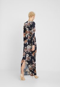 Woman with short curly hair wearing a long black floral dress with a back slit, beige high heels, and dangling earrings, standing against a plain background.