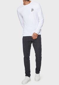 Camisa blanca de manga larga con un logo negro en el pecho, combinada con pantalones de cuadros gris oscuro y zapatillas blancas.