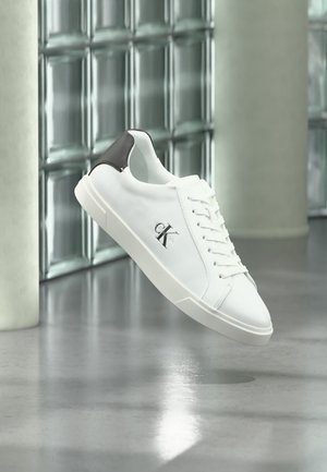 Pantof alb sport cu tab pe călcâi negru și logo „ck”, șiret, prezentat plutind peste podea gri lucioasă, cu perete din blocuri de sticlă și coloane în fundal.