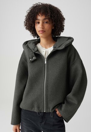Jeune femme aux cheveux bouclés porte une veste zippée gris foncé avec capuche matelassée sur un haut blanc, debout devant un fond clair uni.