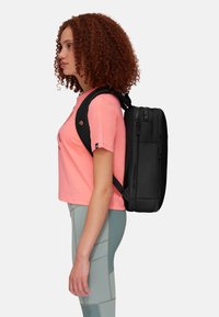 Zaino nero con forma rettangolare, dotato di numerose cerniere e una superficie testurizzata, indossato sopra una maglietta rosa e leggings verde chiaro.