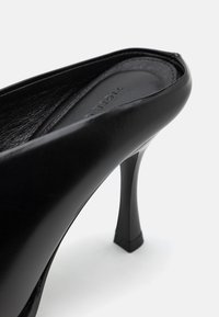 Närbild av en svart läder högklackad sko som visar den böjda klacken och den släta ytan mot en vit bakgrund.