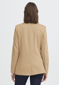 Blazer beige con un design sartorial, maniche lunghe, apertura posteriore e superficie liscia, abbinato a pantaloni in denim scuro.