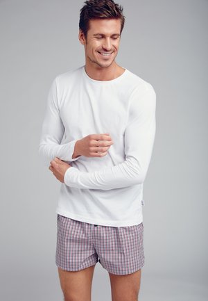 Hvid langærmet skjorte parret med rød- og blåternede boxershorts, der har en afslappet pasform og blødt stof, båret af en smilende mandlig model.