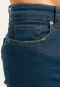 Jeans de mezclilla azul oscuro con un diseño ajustado, que presentan costuras naranjas, dos bolsillos traseros y un borde deshilachado en la parte superior del bolsillo.