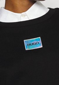 Camicia bianca con colletto sotto un maglione nero, con una patch rettangolare olografica lucente etichettata "HUGO" sul petto.