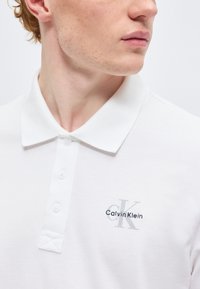 Νέος άνδρας που φοράει λευκό πόλο της Calvin Klein με το λογότυπο στο στήθος, κοιτώντας πλάι σε απλό, φωτεινό φόντο.