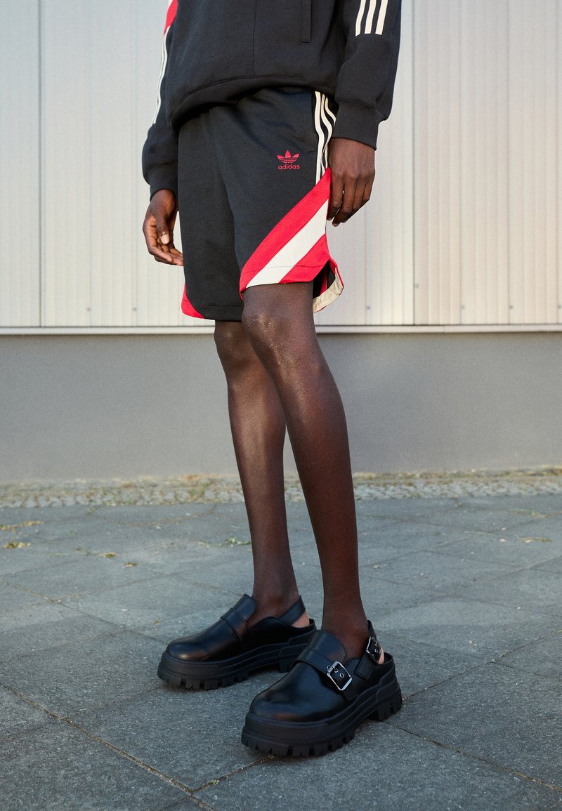 Zapatos de cuero negro con suelas gruesas y detalles de hebilla, combinados con pantalones cortos negros que tienen rayas diagonales rojas y blancas.