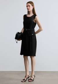 Svart ermeløs kjole med en gjennomsiktig øvre del, innsvingt midjebelte, frontlommer og glatt stoff. Brukt med svarte hæler med stropper.