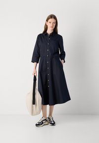 Donkerblauwe blousejurk met lange mouwen, een patchzak en een uitlopende design, gecombineerd met een crèmekleurige tote en gestreepte sneakers.