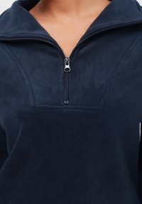 Közeli kép egy szeméről, aki sötétkék fleece pulóvert visel, félig cipzáros magas gallérral, és látható a mellkas és a nyak területe.