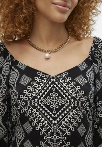 Svart bluse med hvite geometriske mønstre, med pufferm og V-hals. Gullkjede med en stor perle som accent.