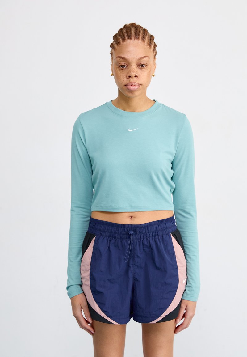Šviesiai mėlynas ilgomis rankovėmis cropped viršus su mažu baltu Nike logotipu, suderintas su tamsiai mėlynomis šortais, turinčiais rožinių ir juodų akcentų bei elastinga juosmens dalimi.