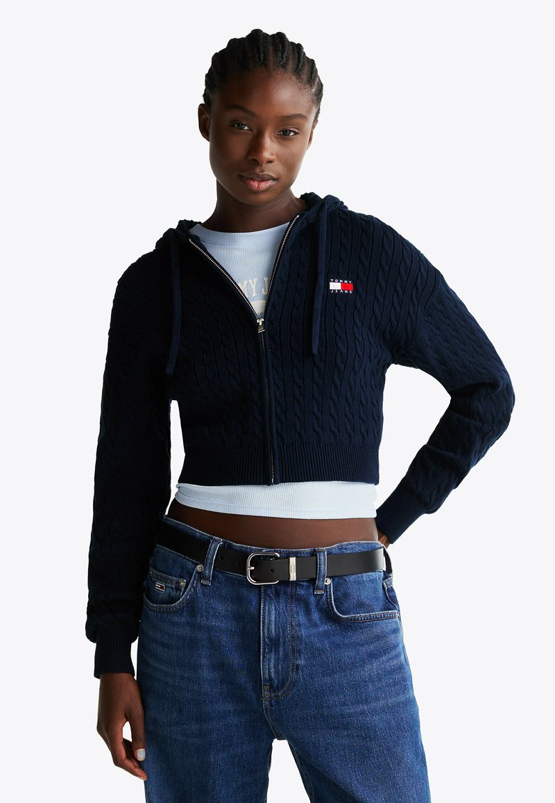 Granatowy sweter z dzianiny w splot warkoczowy, z krótkim krojem, z zamkiem błyskawicznym z przodu, kapturem ze sznurkiem i małą naszywką z logo. W komplecie z jasnoniebieską bluzką i dżinsami.