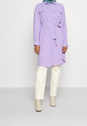 Kvinde iført en lavendelfarvet skjortekjole med bælte over hvide bukser og beige ankelstøvler mod en ensfarvet lys baggrund.