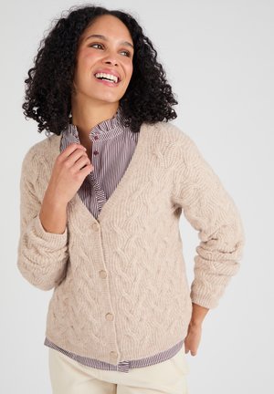 Donna sorridente con capelli ricci indossa un cardigan a trecce beige sopra una camicia a righe con bottoni e pantaloni chiari, posando con una mano vicino al colletto.