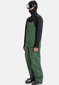 Persoon in donkergroene bretelsneeuwbroek, zwart longsleeve top, zwarte beanie, skibril en zwarte skibotten, staande tegen een witte achtergrond.