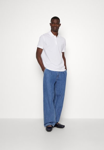 Camisa polo blanca, pantalones vaqueros azules holgados, mocasines negros. La polo tiene cuello y mangas cortas; los pantalones tienen un diseño de corte recto.