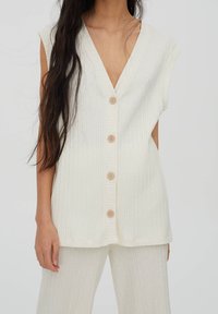 Top sin mangas color blanco roto con un profundo escote en V, textura acanalada y cinco botones de madera en la parte delantera, combinado con pantalones acanalados a juego.