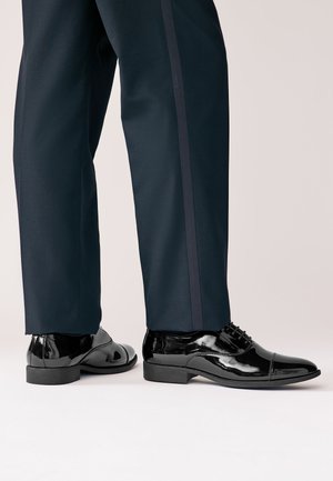 Glänzende schwarze Halbschuhe, getragen zu dunkelblauen Hosen mit einem dezenten Seitenstreifen, von den Unterschenkeln abwärts gezeigt.