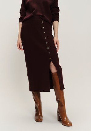 Mujer con falda midi marrón oscuro con botones laterales y una abertura frontal, combinada con botas altas de cuero marrón con tacón.
