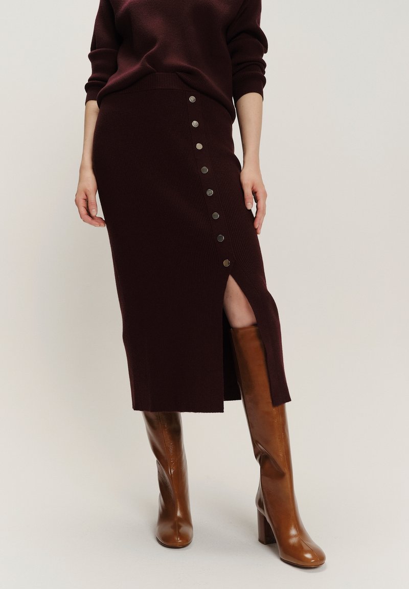 Femme portant une jupe midi marron foncé avec des boutons latéraux et une fente à l'avant, associée à des bottes hautes en cuir marron à talons.