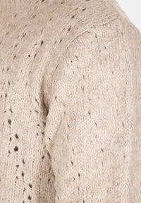 Maglione beige lavorato a maglia con un motivo testurizzato, caratterizzato da piccole macchie scure, scollatura tonda e dettagli di cucitura ben visibili sulla spalla.