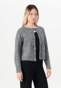 Mladá žena v šedej pletenej cardigan s veľkými gombíkmi, oblečená v čiernom topu a čiernych nohaviciach, postávajúca na hladkom bielom pozadí.