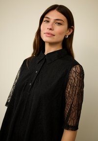 Melnas mežģīņu blūze ar apkaklīti, īsām mežģīņu piedurknēm un vaļēju piegriezumu ar pogu detaļām priekšpusē.