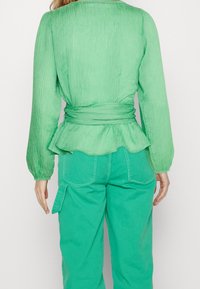 Zelená texturovaná blůza s dlouhými rukávy, peplum pasem a zavázaným páskem, spárovaná s odpovídajícími zelenými kalhotami s širokými nohavicemi a bočními kapsami.
