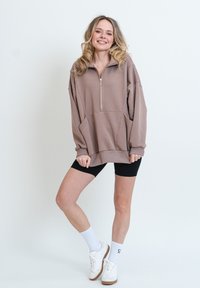 Pullover in tinta marrone chiaro, oversize, con zip a un quarto, tasche frontali e polsini a coste, abbinato a pantaloni corti neri e sneakers bianche.