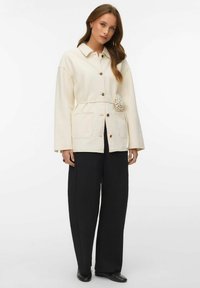 Naine, kes kannab kreemikollast nööbitavat jopet, millel on vöökoha lilledetail, musti laiade säärtega pükse ja musti lameda tallaga kingi, seisab valge tausta ees.