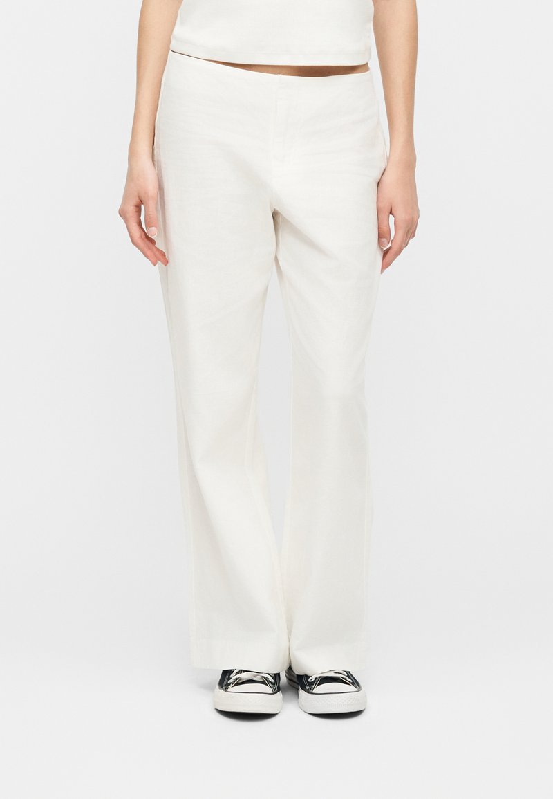 Άτομο που φορά λευκό παντελόνι με φαρδύ πόδι, ασπρόμαυρα αθλητικά παπούτσια, και κοντό λευκό μπλουζάκι, στέκεται μπροστά σε απλό φόντο.