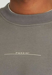 Sweat-shirt gris avec un col côtelé, portant le nom de la marque "CHASIN’" en blanc au-dessus d'une fine ligne horizontale. Texture douce.