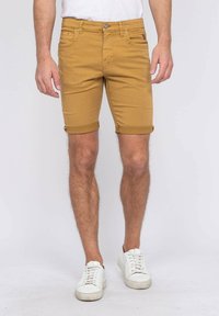 Shorts jaune moutarde en tissu coton, avec des poignets roulés, des passants de ceinture et des poches avant. Associés à des baskets blanches.