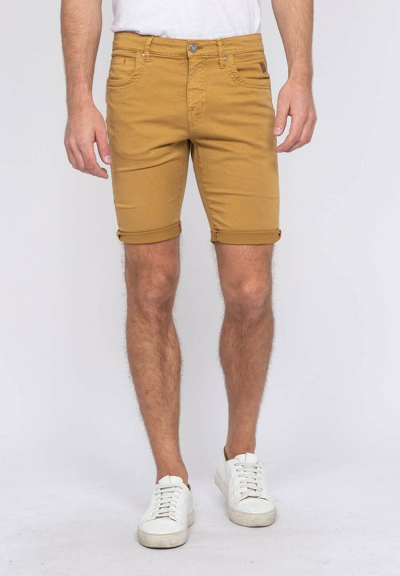 Shorts jaune moutarde en tissu coton, avec des poignets roulés, des passants de ceinture et des poches avant. Associés à des baskets blanches.