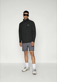 Sort langærmet trøje med halv lynlås, grå shorts og sportssko. Solbriller på. Casual udendørsbeklædning med Patagonia-logo på brystet.