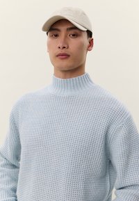 Mężczyzna w beżowej czapce i jasnoniebieskim teksturowanym swetrze z golfem, stojący na tle jednolitego, jasnokremowego tła.