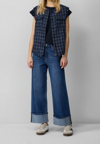Blå tweedvest med store forlommer, lagd ovenpå en sort top. Sammen med brede denimjeans med foldede manchetter og hvide sneakers.