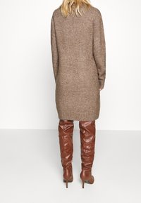 Robe pull longue en maille marron avec une coupe ample, une texture côtelée et des fentes sur les côtés, associée à des bottes stiletto hautes en cuir marron.