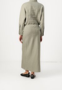 Robe longue en lin vert olive avec des poignets à boutons et un col en V discret, associée à des chaussures plates noires ornées d'un détail rond.