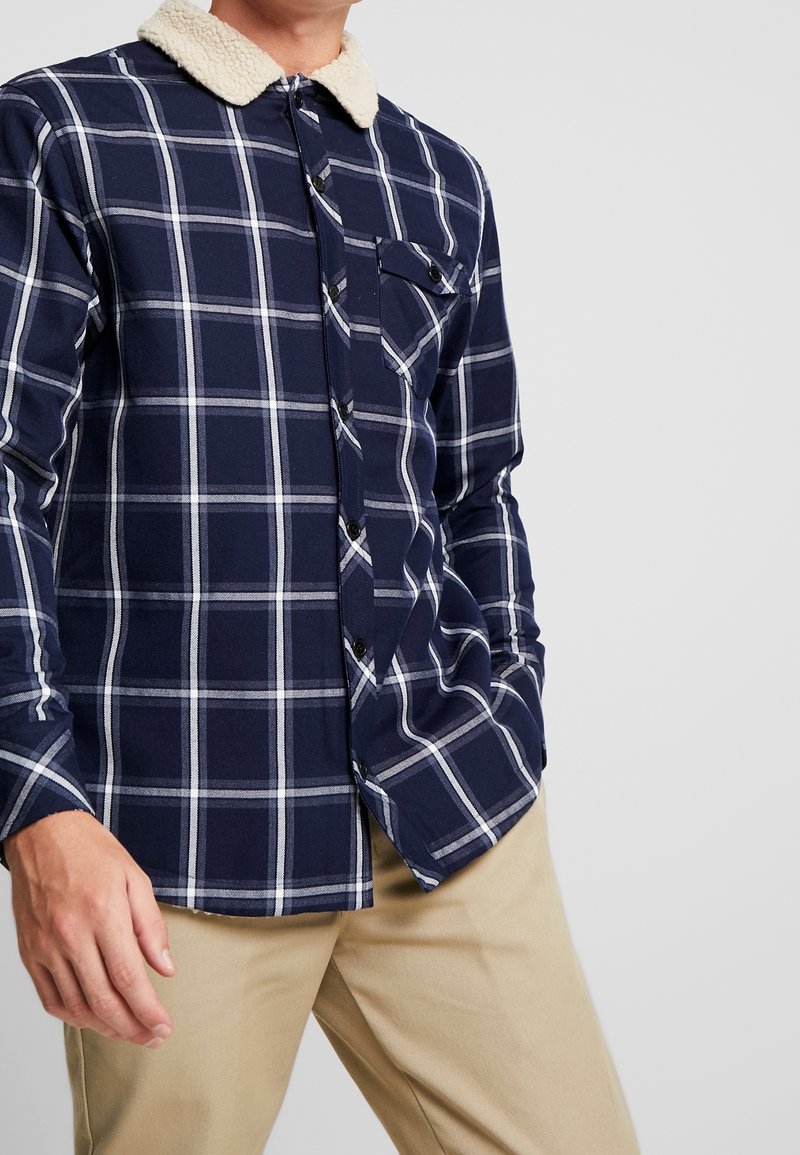 Homme portant une chemise à carreaux bleu marine avec col en polaire blanche et pantalon beige, debout avec une main dans la poche.